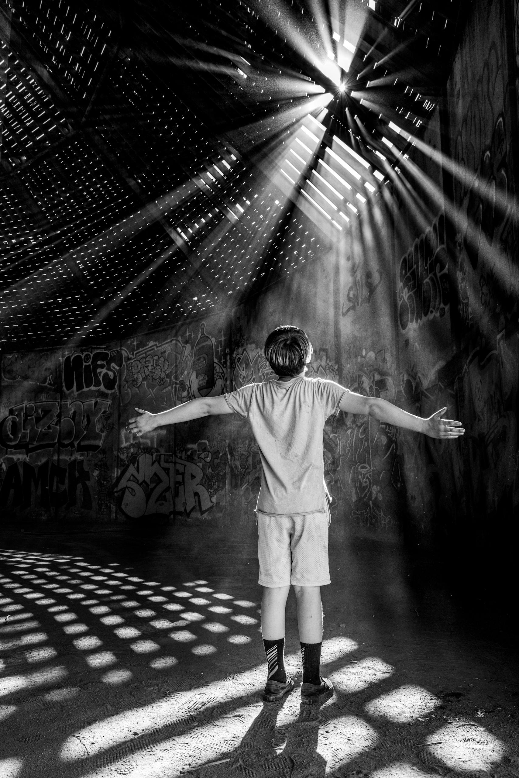 A boy stands with open arms in dramatic light from roof slats in an abandoned graffiti-covered building.
